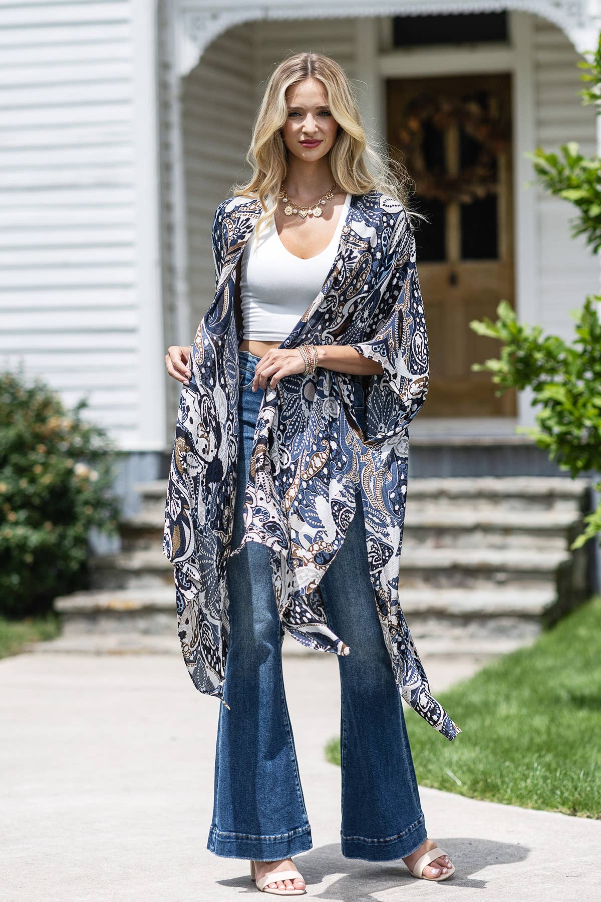 Woman wearing a patterned kimono over a white top and blue jeans in front of a house.
