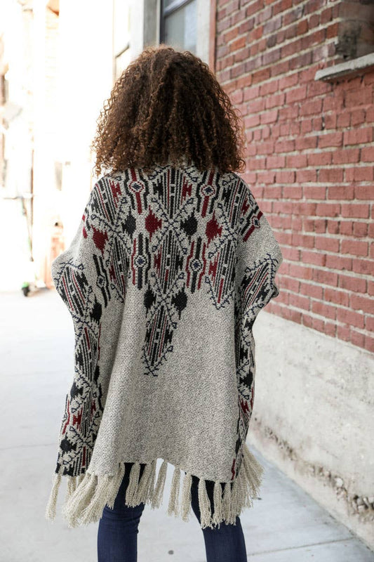 Person wearing a patterned cardigan with fringe, standing in front of a brick wall.
