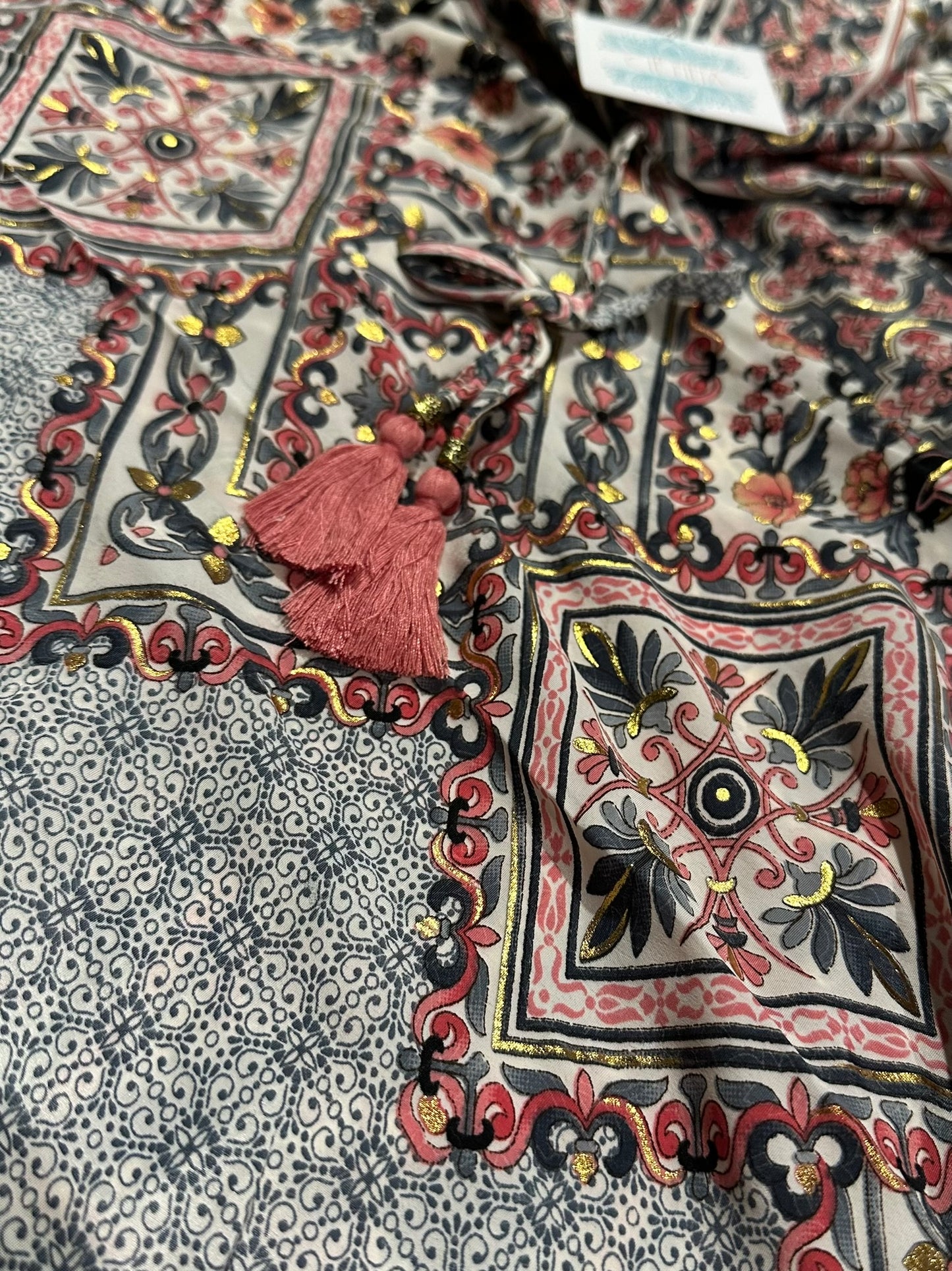 A close-up of a printed fabric with a floral and geometric pattern in red, black, beige, and yellow, featuring a prominent dusty pink tassel ornament.