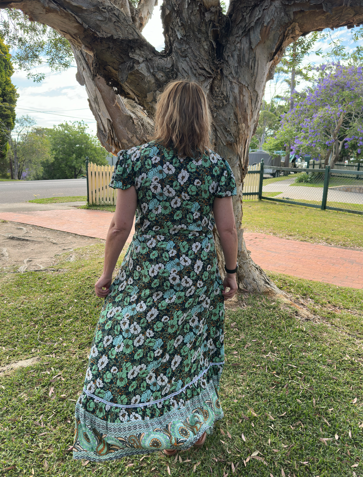 Person wearing a floral dress standing under a large tree in a park-like setting.