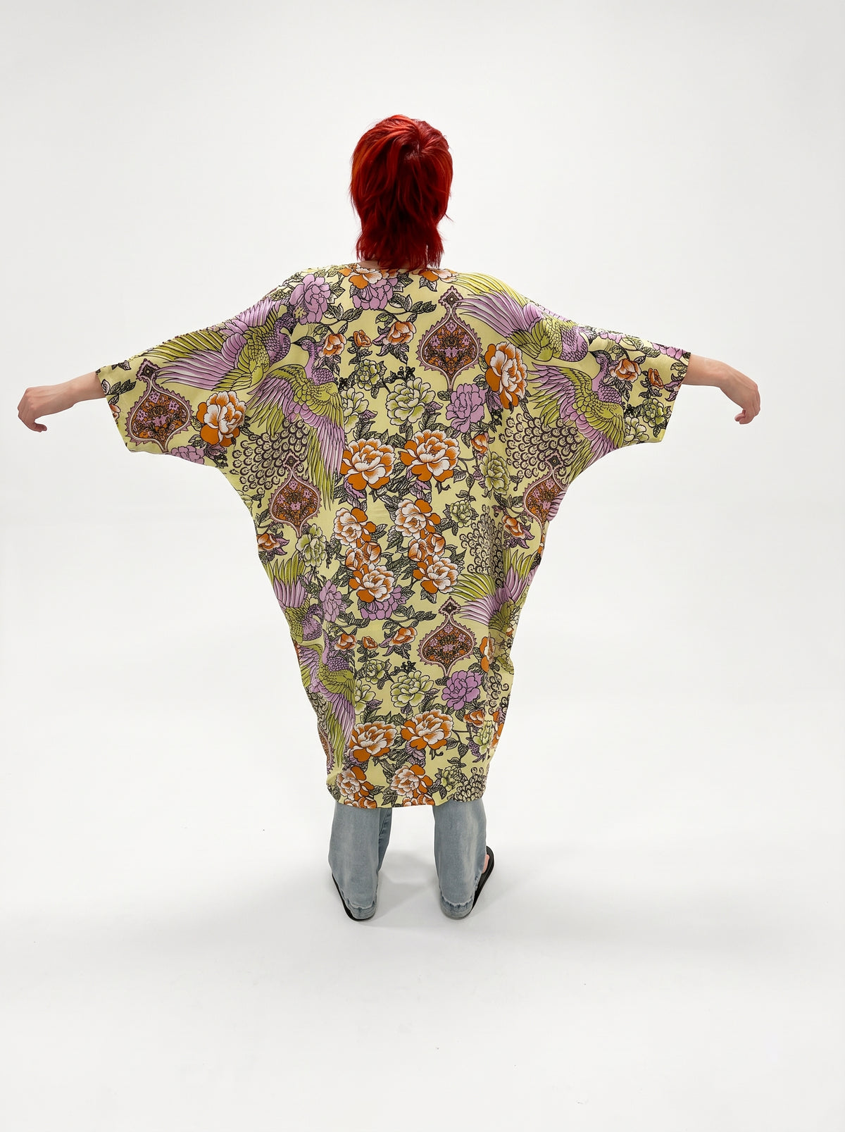 A person viewed from behind wearing a loose, long-sleeved kaftan-style top with a dense floral and botanical print in yellow, purple, orange, and green, paired with gray pants, against a plain white studio background.