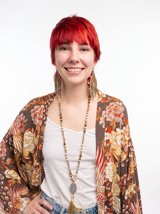 A smiling woman with short bright red hair is shown from the chest up against a plain white studio background. She wears a white tank top, blue jeans, long dangling earrings, a long beaded necklace, and a loose brown floral kimono-style cardigan.