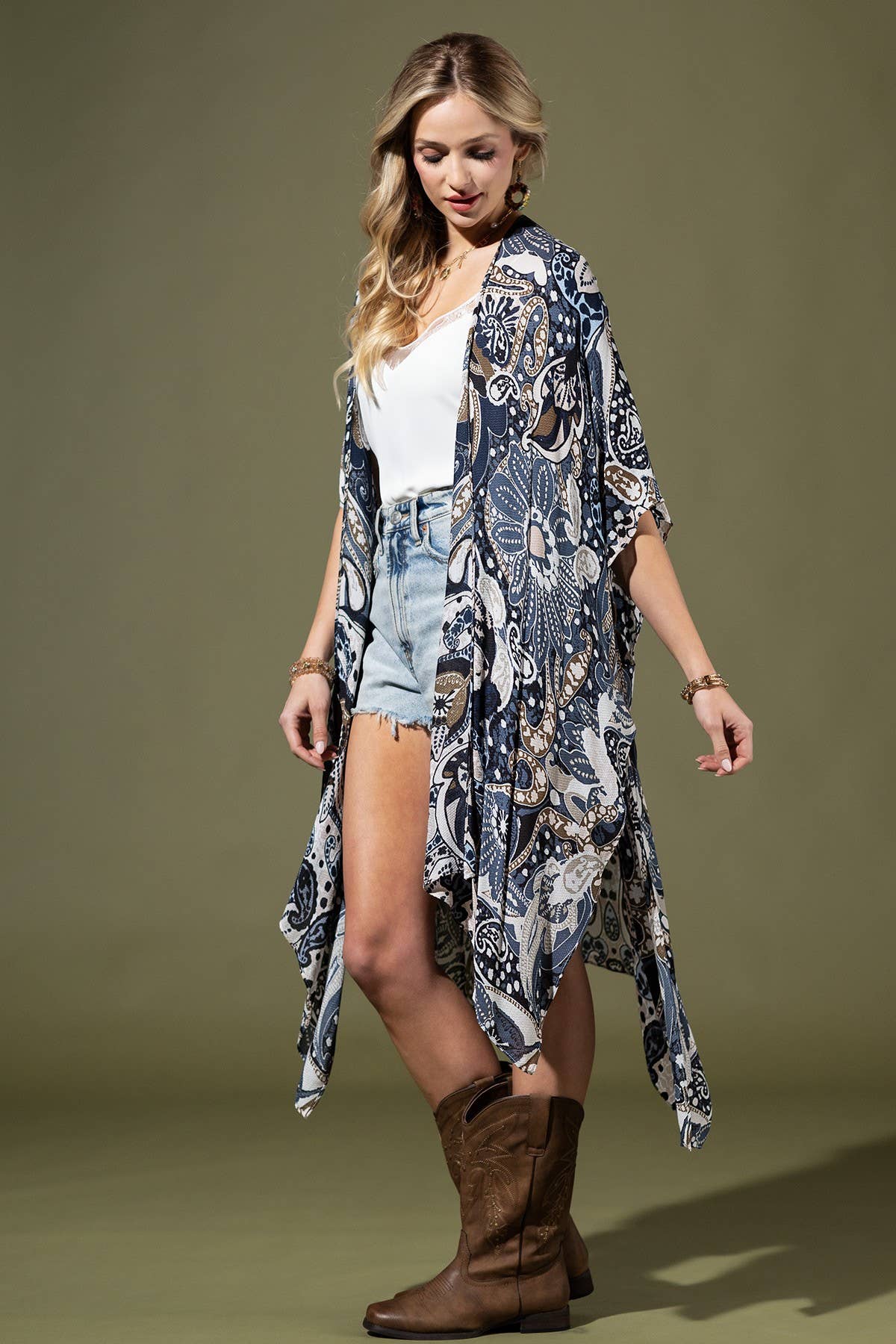 Woman wearing a patterned kimono, white top, denim shorts, and brown boots on a green background
