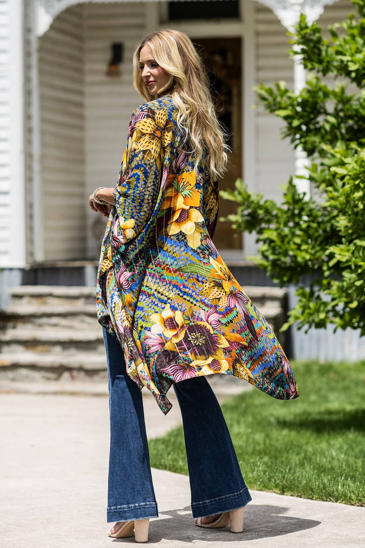 Woman wearing a colorful floral coat and blue jeans standing in front of a house.