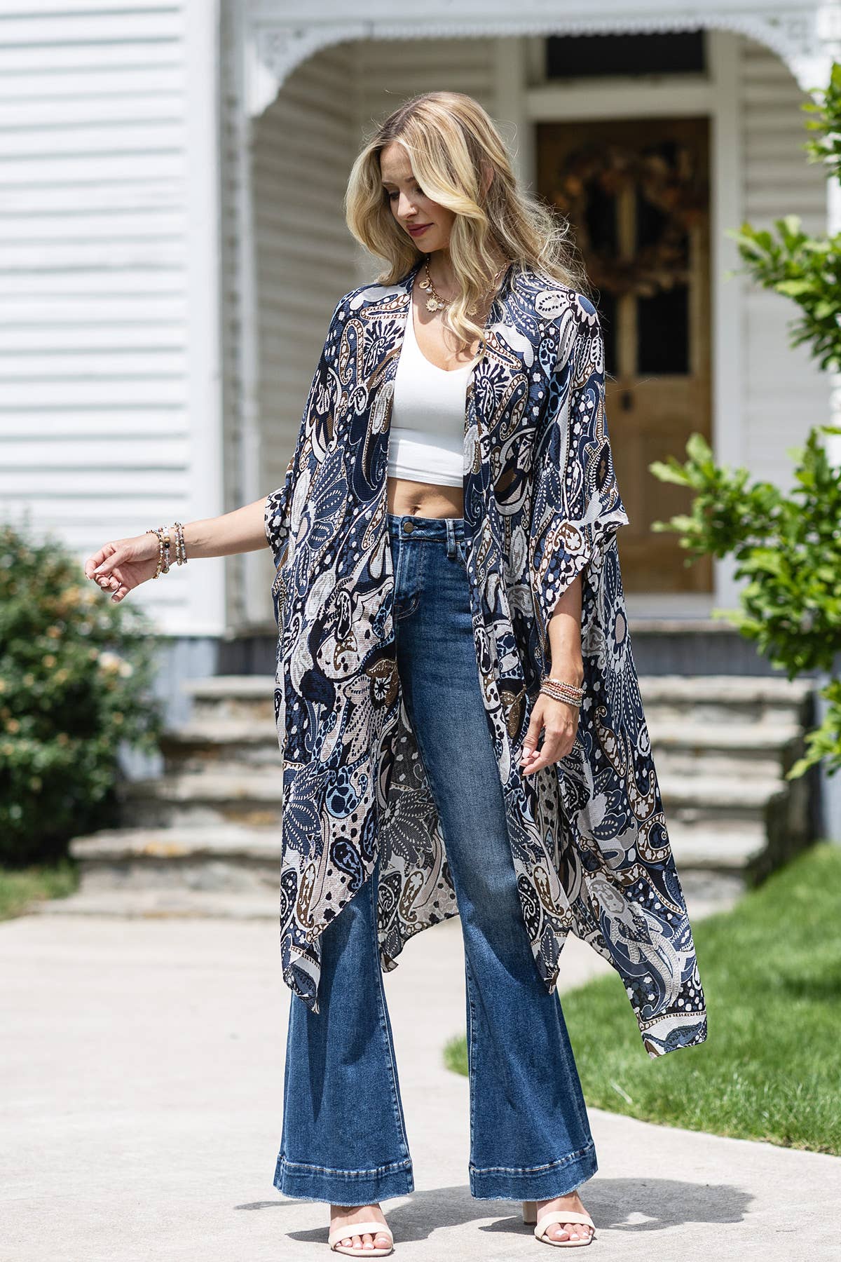 Woman wearing a floral kimono over a white top and blue jeans, standing outside a house.