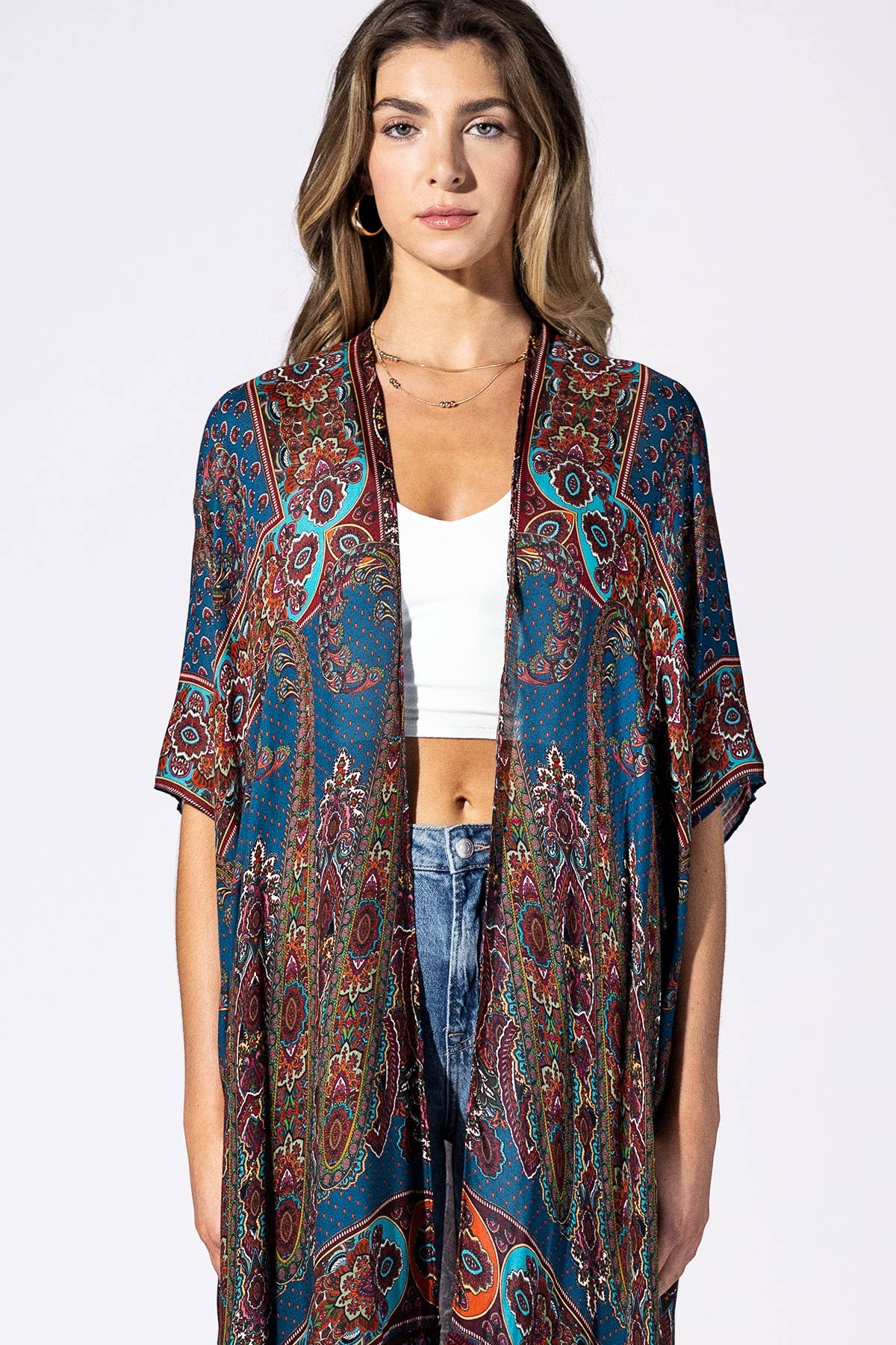 A woman modeling a long-sleeve open-front kimono cardigan with a vibrant paisley pattern in teal, rust red, and brown over a white crop top and blue jeans, shot against a solid white studio background.