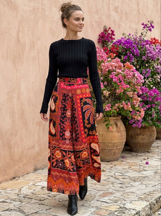 Woman wearing a black top and patterned skirt standing in front of a stone wall with flowers.