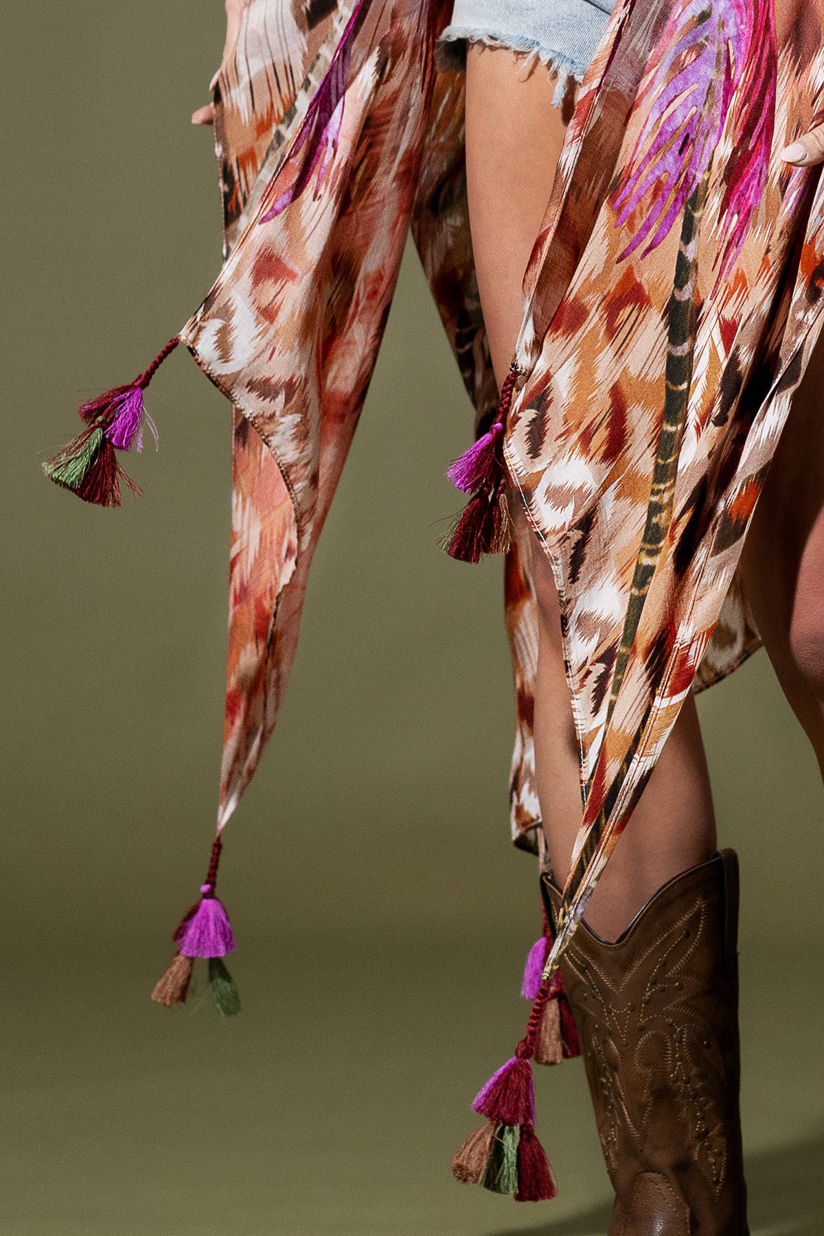 Person wearing a colorful patterned scarf with tassels and cowboy boots on a green background