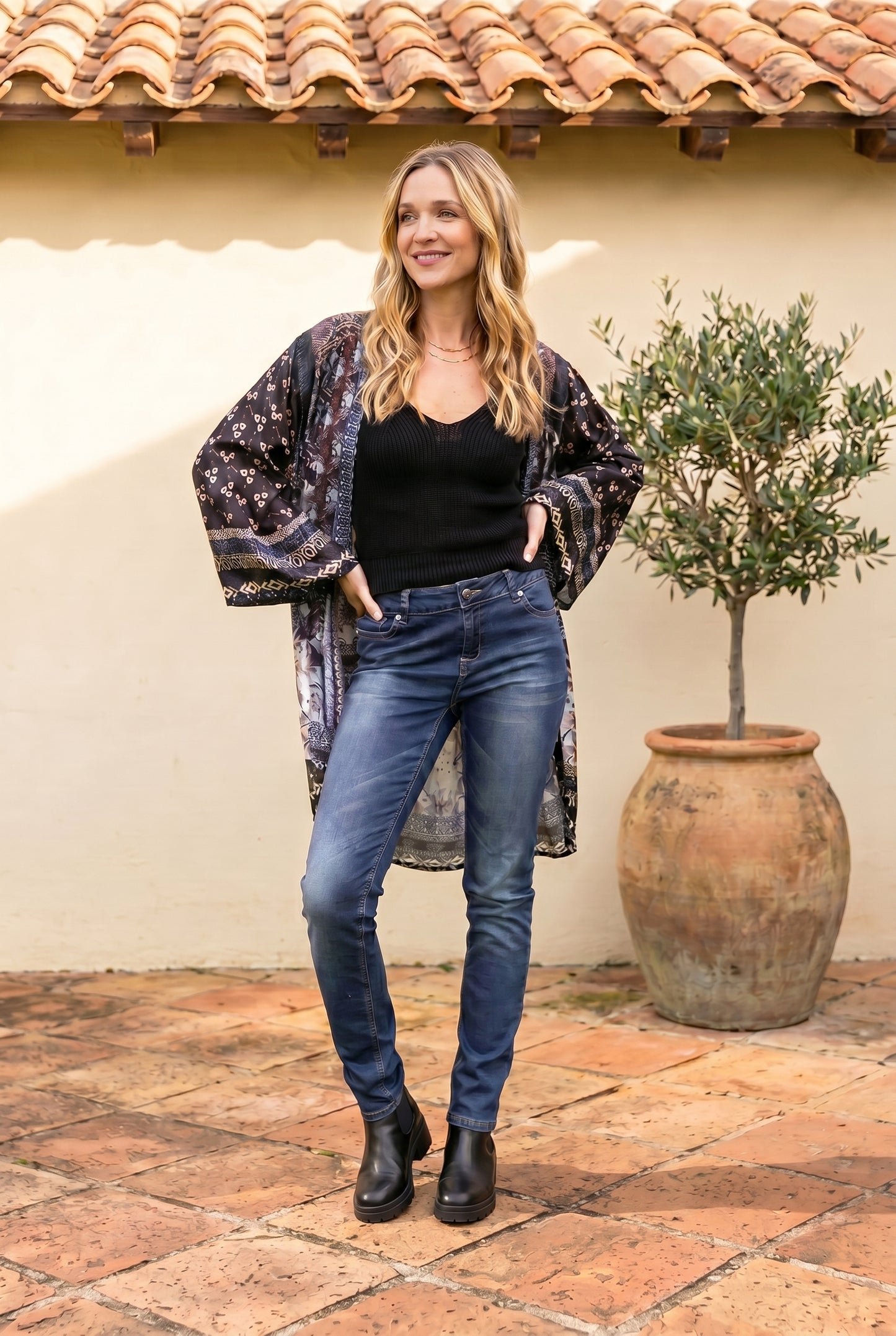 Woman wearing a floral kimono, black top, and blue jeans standing in front of a textured wall with a potted plant.
