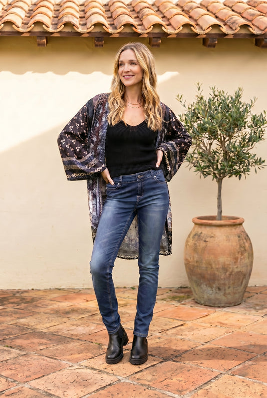 Woman wearing a floral kimono, black top, and blue jeans standing in front of a textured wall with a potted plant.