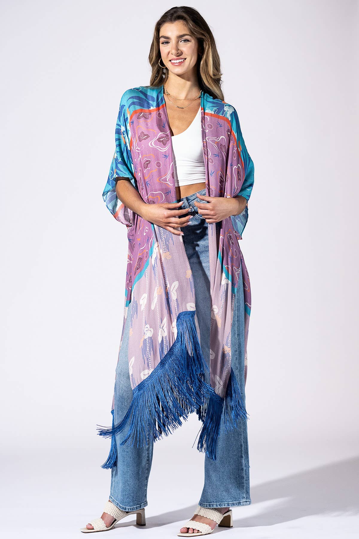 A woman models a long, open-front kimono-style cover-up with wide sleeves and blue fringe, featuring a multicolor paisley and abstract print in purple, blue, pink, and orange. She is styled with blue jeans, a white crop top, and white heeled sandals against a plain white studio background.