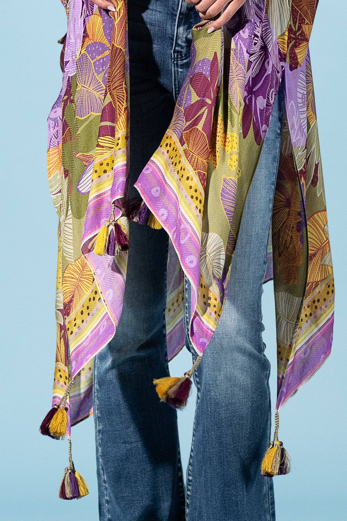 Colorful patterned scarf with tassels draped over a person wearing jeans on a light blue background
