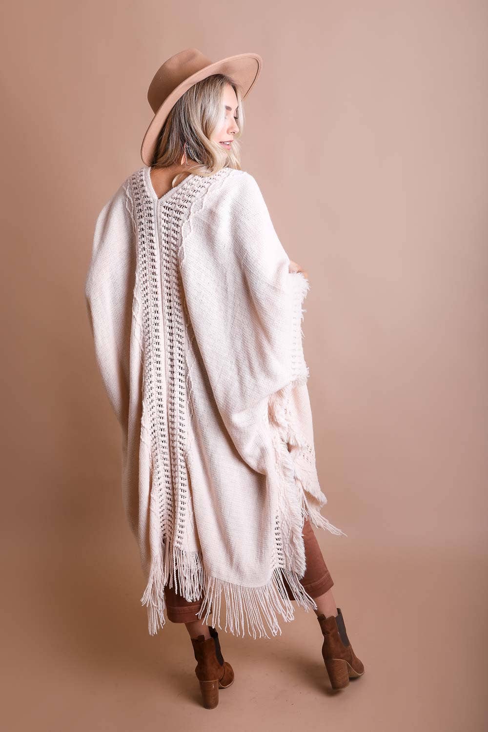 Woman wearing a long, light-colored cardigan with fringe details on a beige background