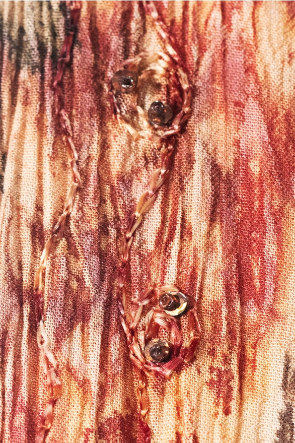 Close-up of a textured fabric with a distressed, splattered pattern in shades of red, brown, and beige, featuring three decorative metal rivet embellishments.