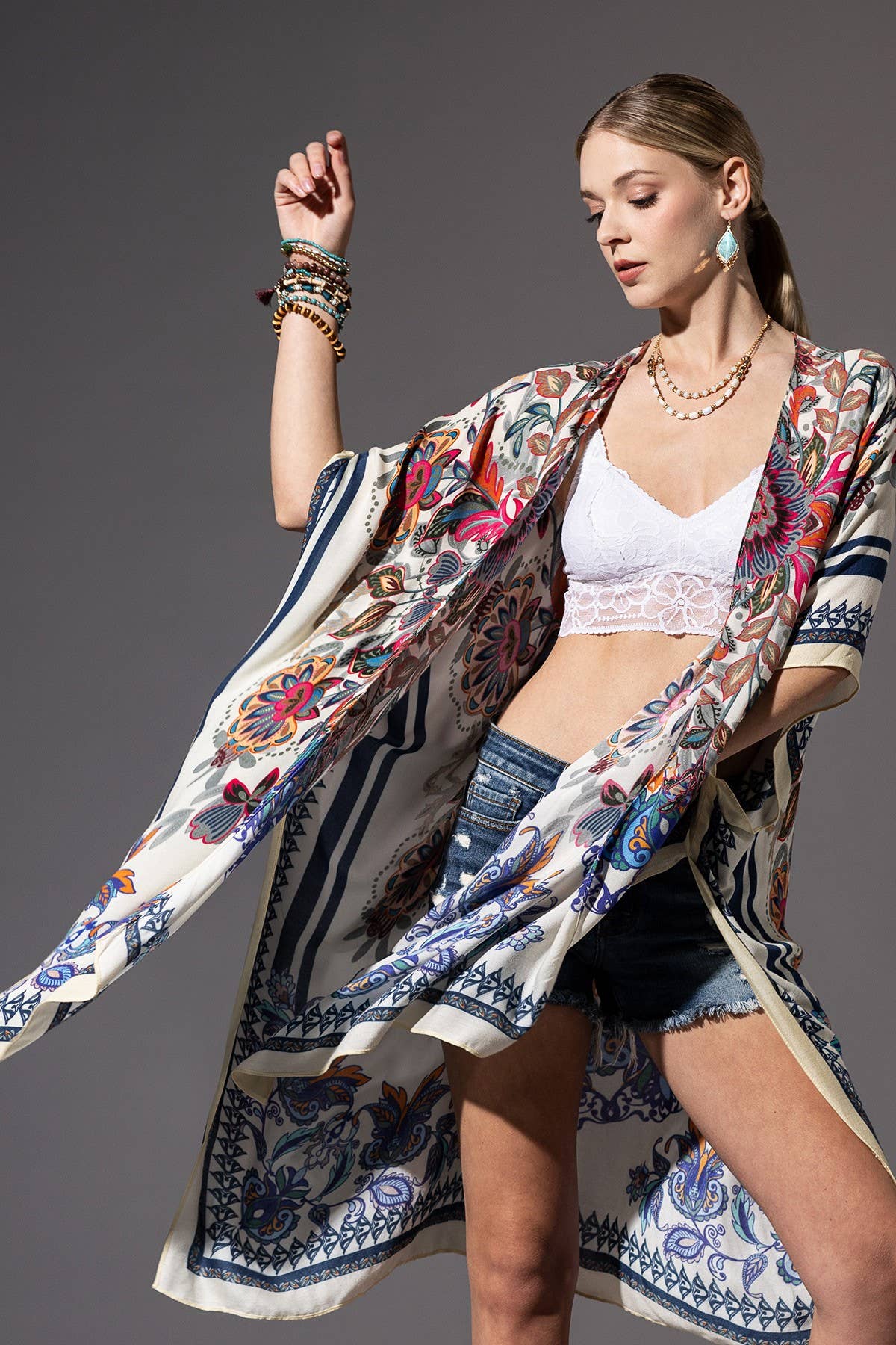 Woman wearing a colorful kimono-style jacket over a white crop top and denim shorts on a gray background