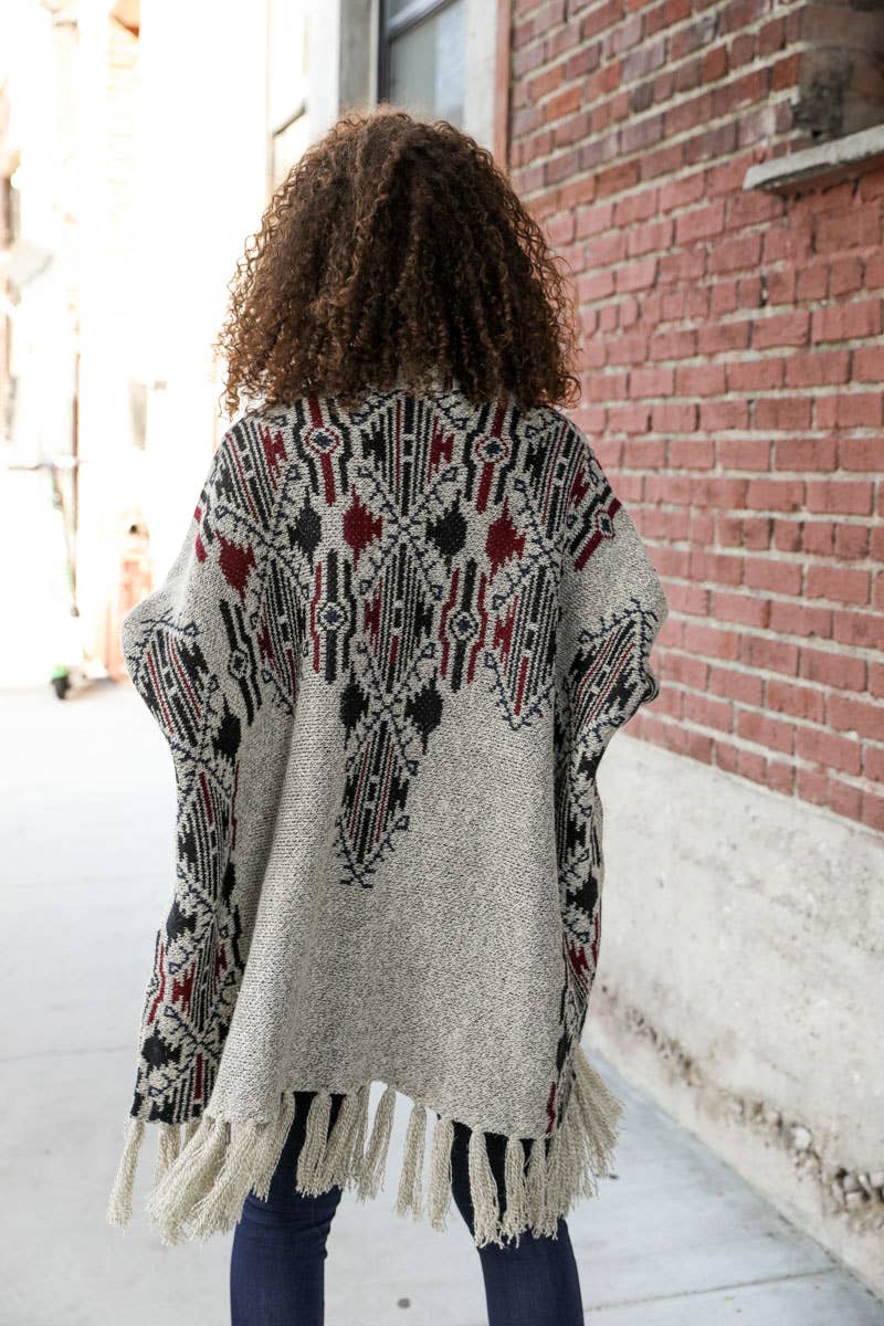 Person wearing a patterned cardigan with fringe, standing in front of a brick wall.