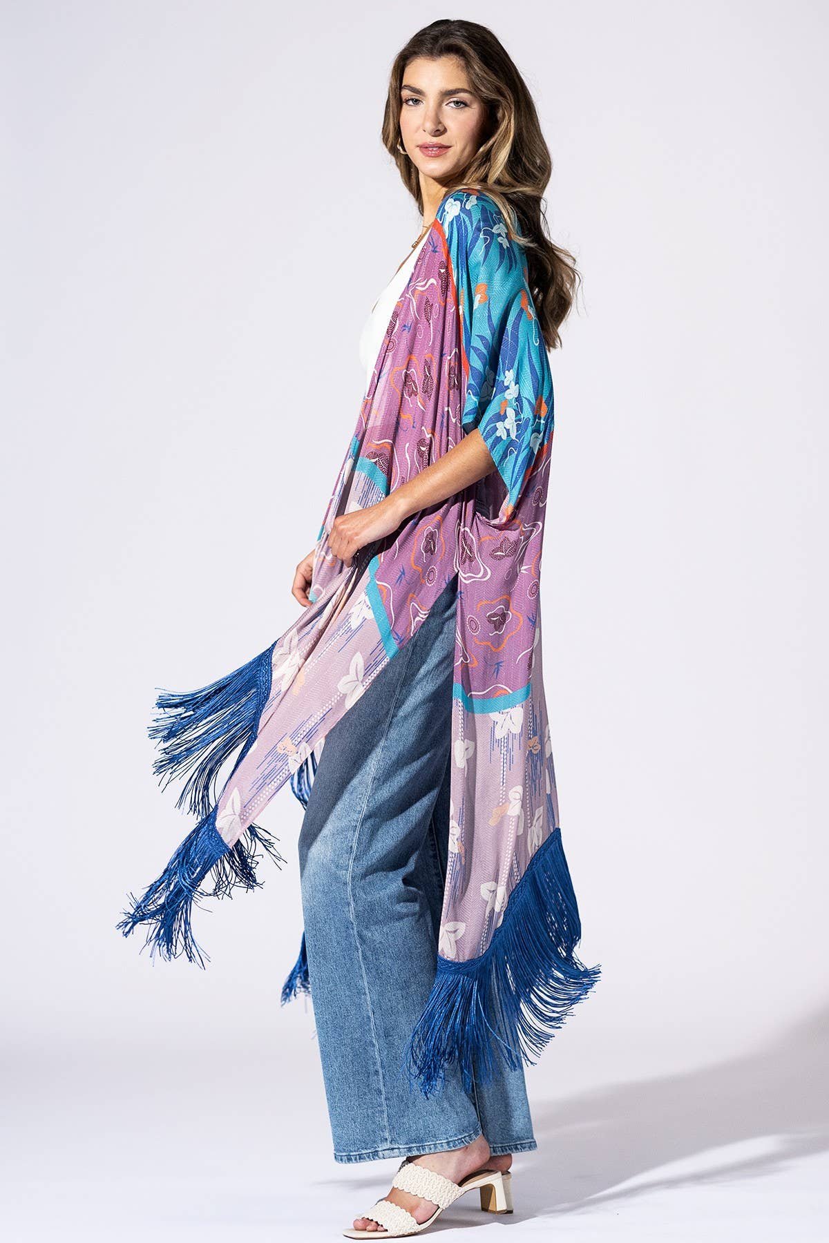 A woman modeling a long, lightweight printed shawl or wrap with blue fringe, worn open over a white top and blue jeans against a plain light gray studio background.