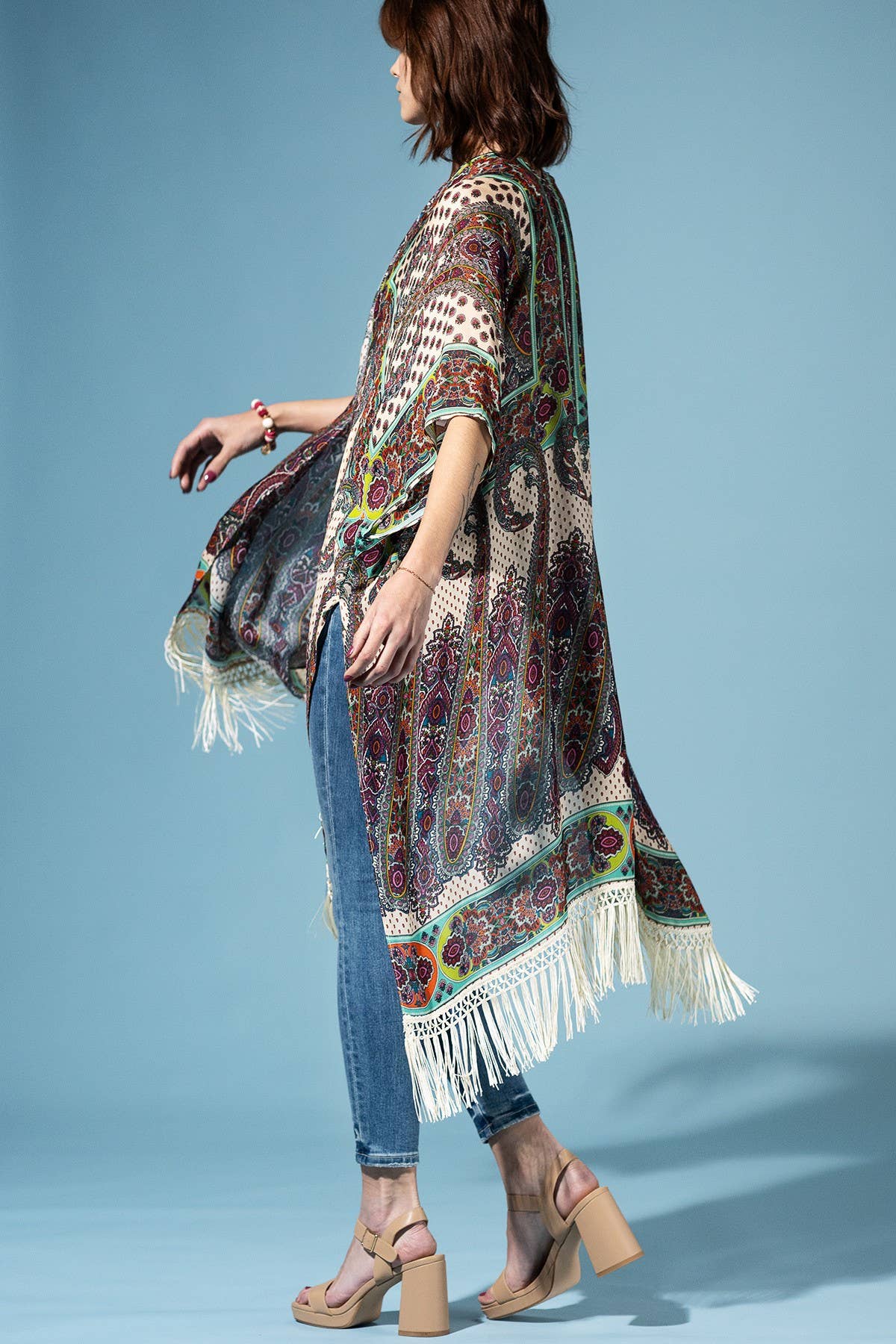 Woman wearing a patterned kimono with fringe over jeans on a blue background