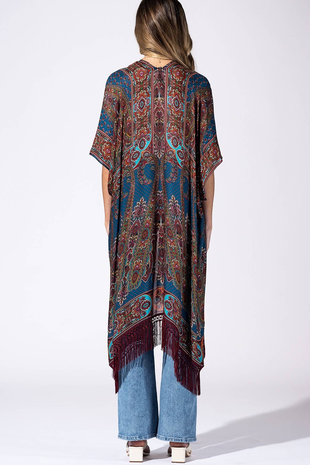 A woman viewed from the back wearing a loose, short-sleeve patterned poncho or tunic with fringe at the hem, paired with light blue jeans and white block-heel sandals, against a plain white studio background.