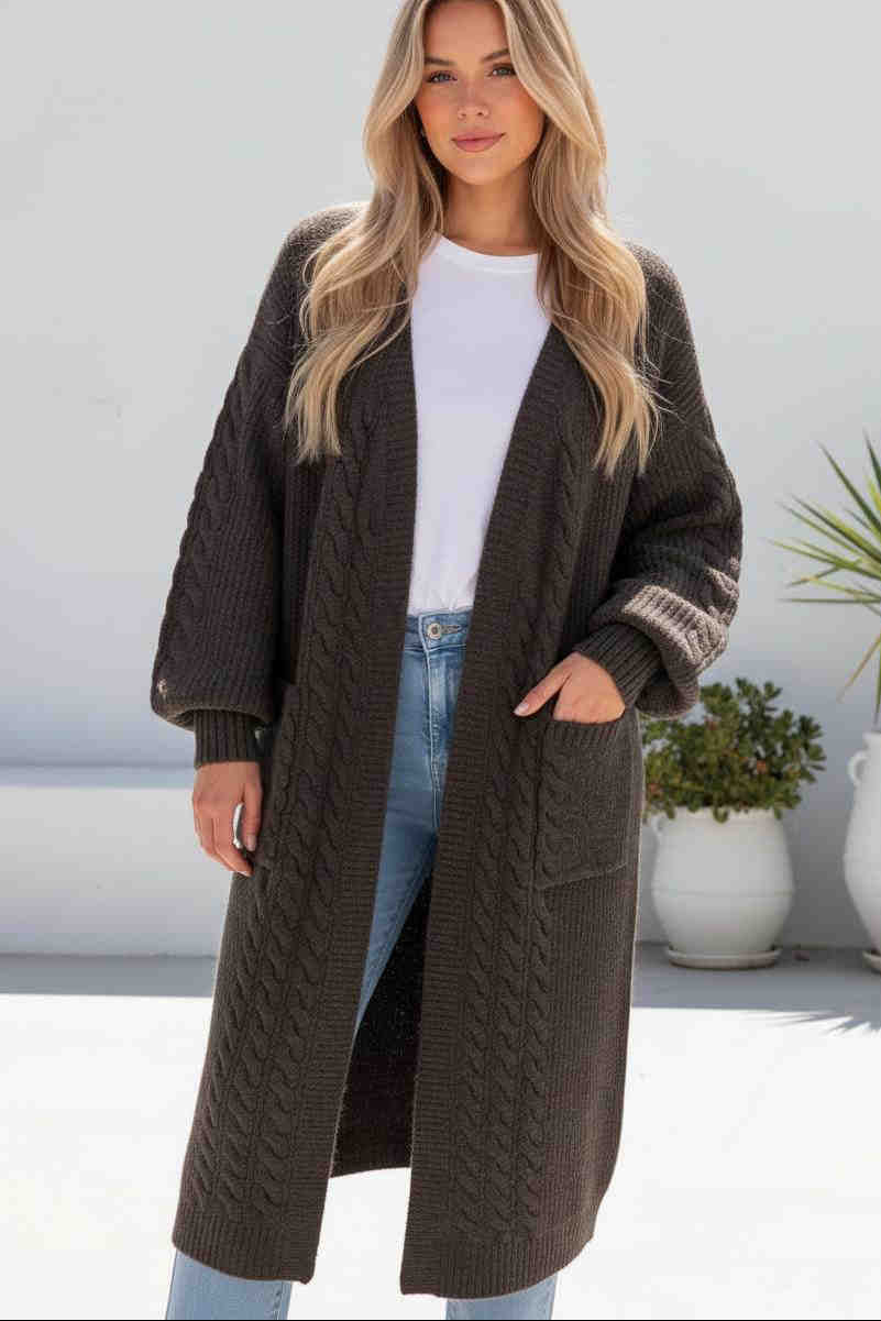 Woman wearing a long brown knit cardigan with a white top and blue jeans, standing against a light background.