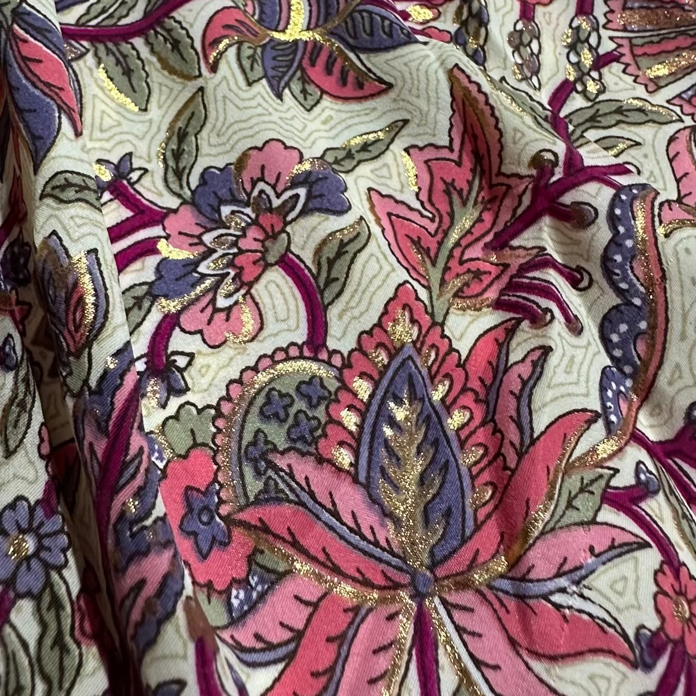 Close-up of a fabric with intricate floral and paisley pattern in pink, purple, and gold on a beige background.