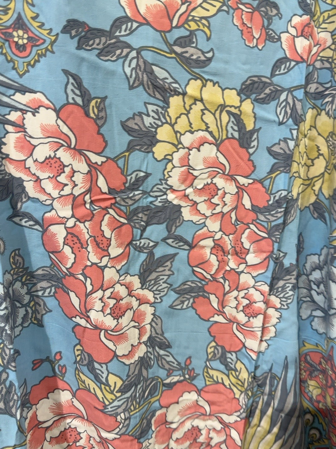 A close-up of a floral fabric print featuring large peony-like flowers in coral red, cream, and yellow with gray leaves on a dusty blue background.