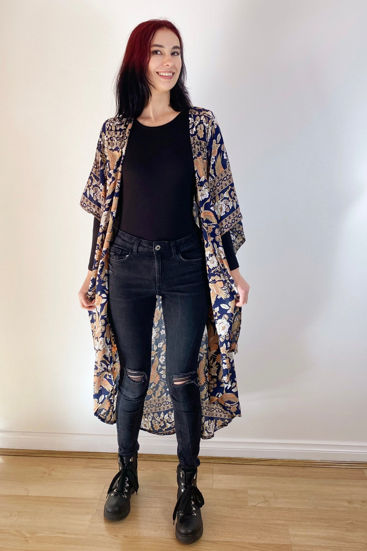 Person wearing a long, patterned cardigan over a black top and jeans, standing against a plain white wall.