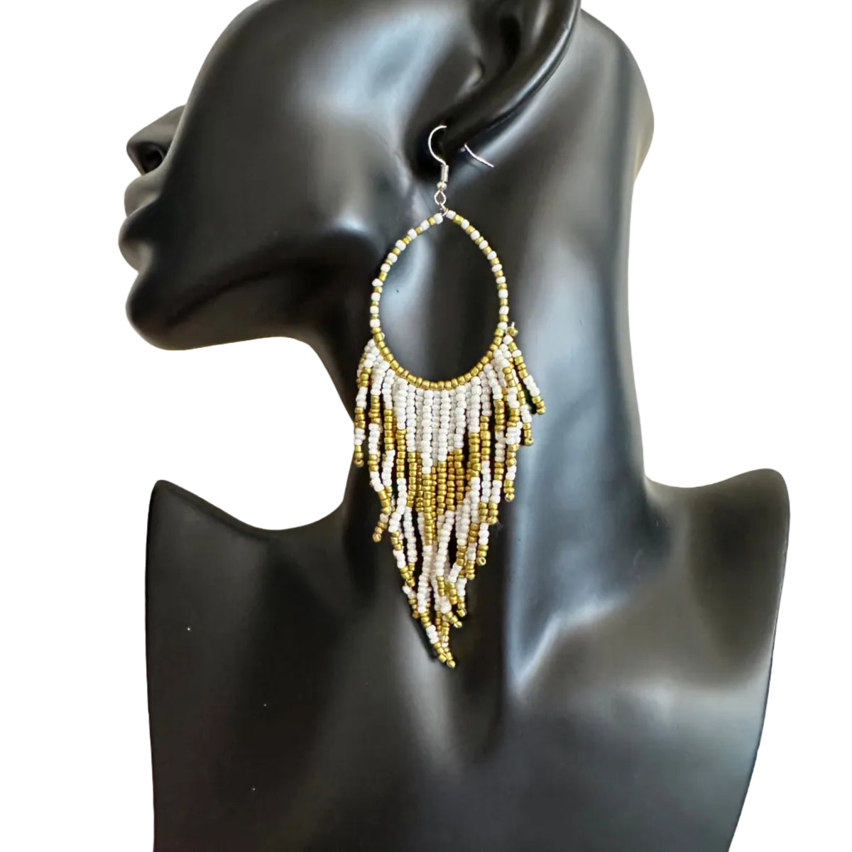 A pair of large hoop earrings with dangling fringe made of white and gold beads, displayed on a black mannequin bust against a beige wall.