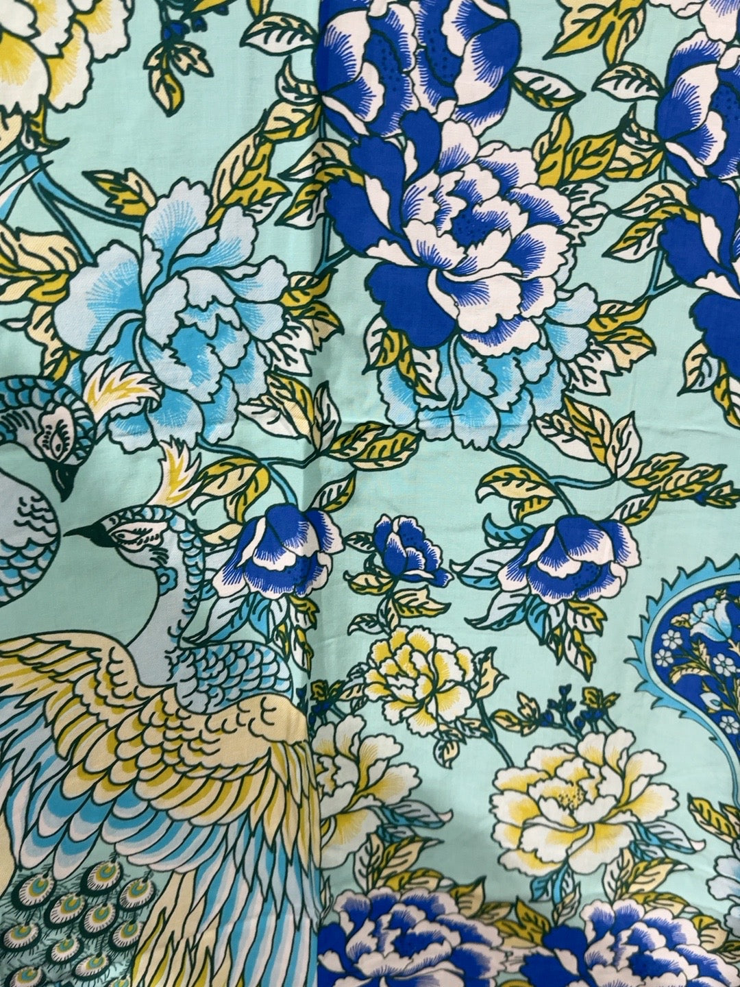 A close-up of a printed textile featuring large blue and white peonies, smaller yellow flowers, and a peacock with blue, yellow, and white feathers on a pale aqua background.