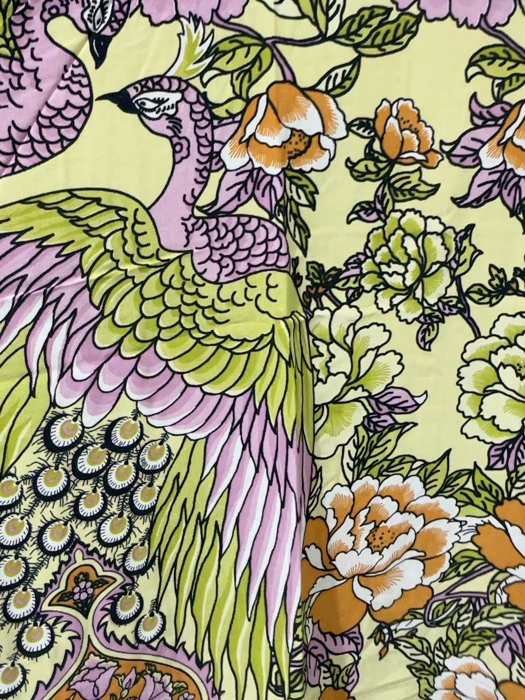 A close-up of a printed textile or wallpaper pattern featuring a large peacock in pink, green, and yellow tones alongside floral elements on a pale yellow background.