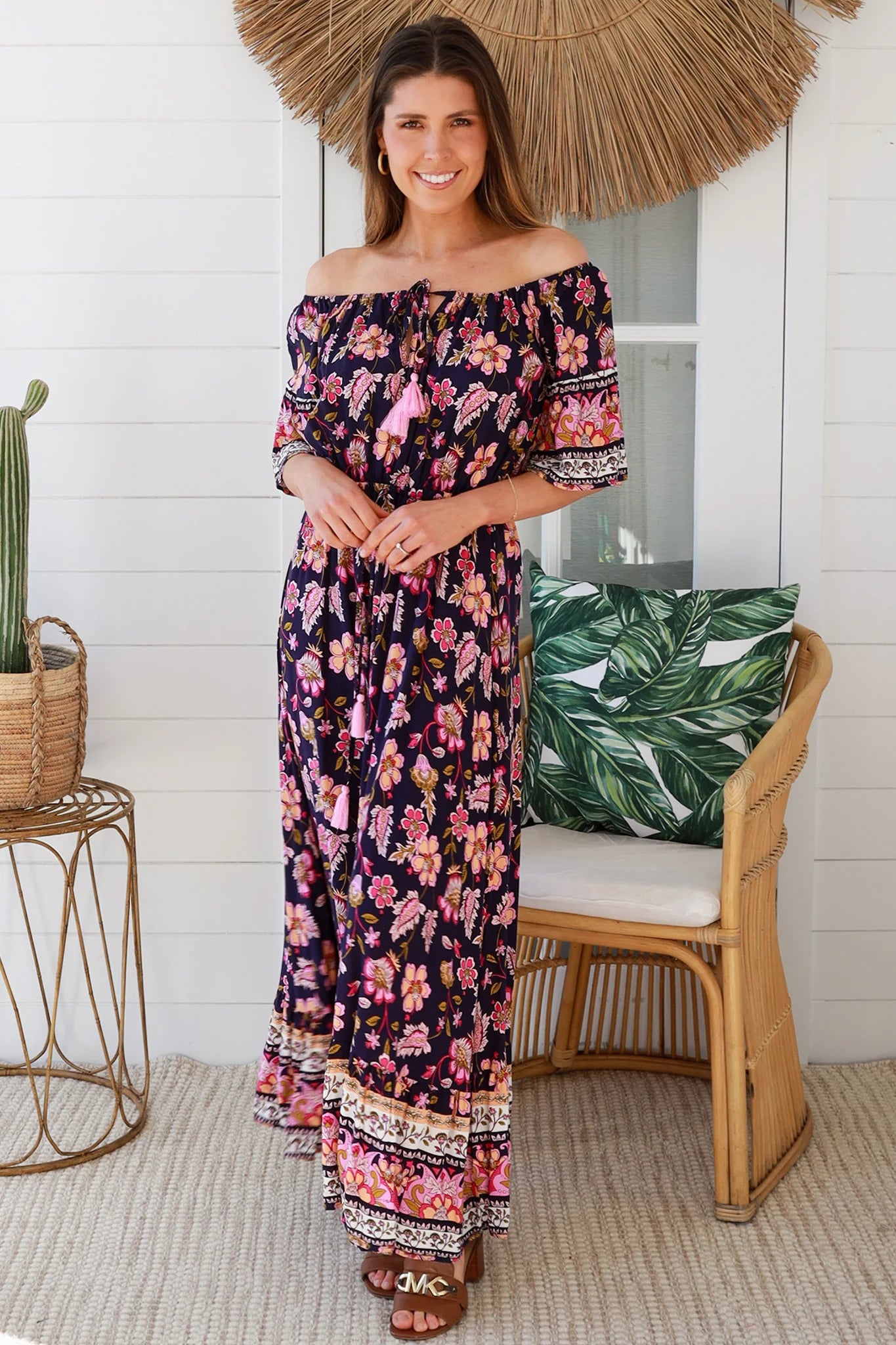 Woman in a floral off-shoulder dress standing in a room with a chair and decorative items.