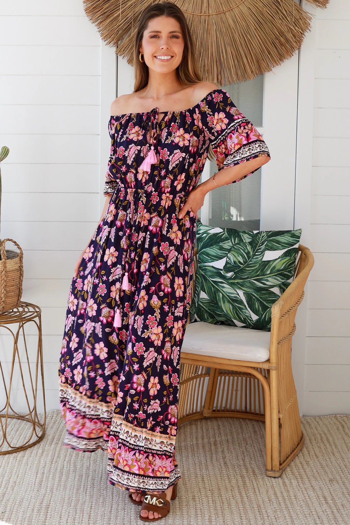 Woman in a floral off-shoulder dress standing in a bright room with a chair and decorative items.