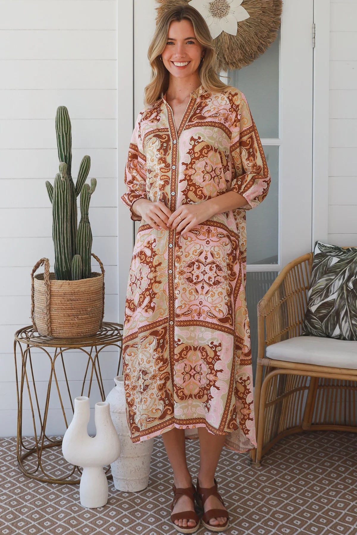 A woman modeling a bohemian-style long kaftan dress with a pink, rust, and beige floral pattern, standing on a porch next to a cactus and a wicker chair.