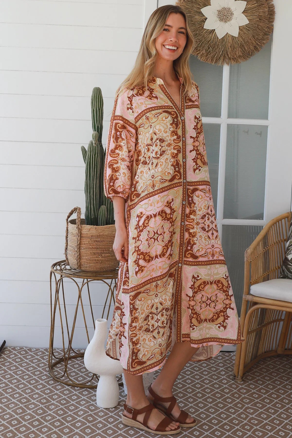 A woman modeling a long-sleeve bohemian-style maxi dress with a pink, brown, and beige patchwork print, standing on a patterned porch floor next to a cactus in a woven basket.