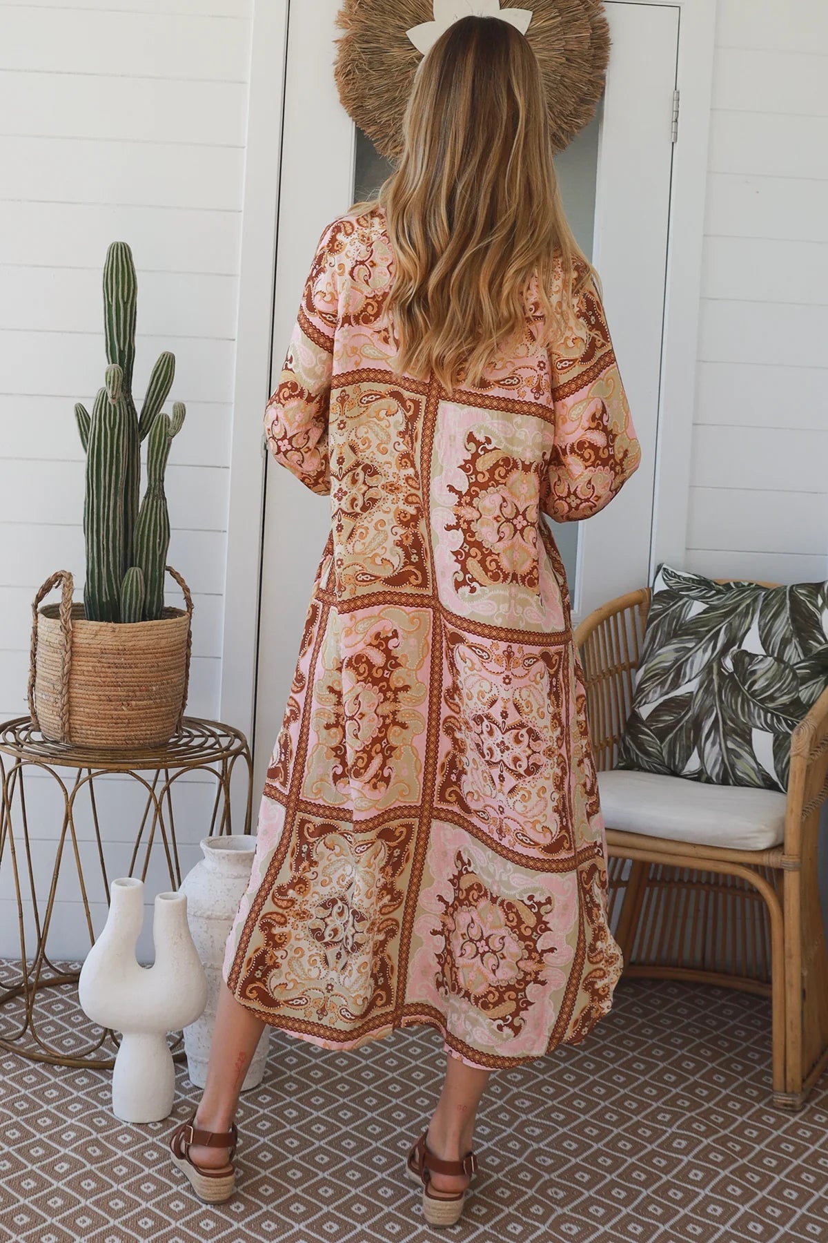 A woman viewed from behind wearing a long patchwork maxi dress with bohemian floral and mandala patterns in pink, beige, and brown. She is standing in a bright, modern porch setting with a cactus, a chair, and a patterned rug.