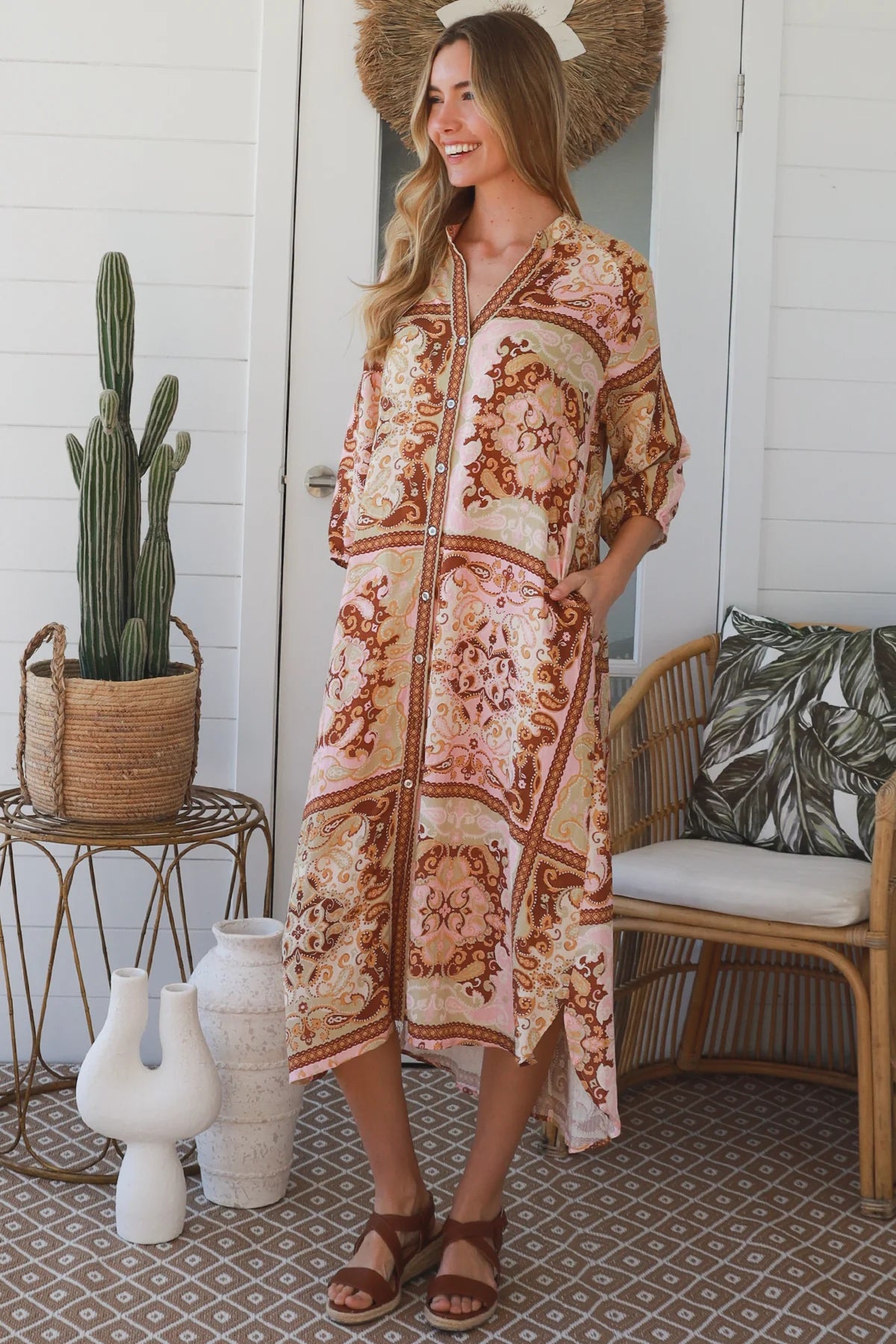A woman wearing a bohemian-style midi kaftan dress with a brown and beige paisley pattern, standing on a patterned porch with a cactus, wicker chair, and decorative vases in the background.