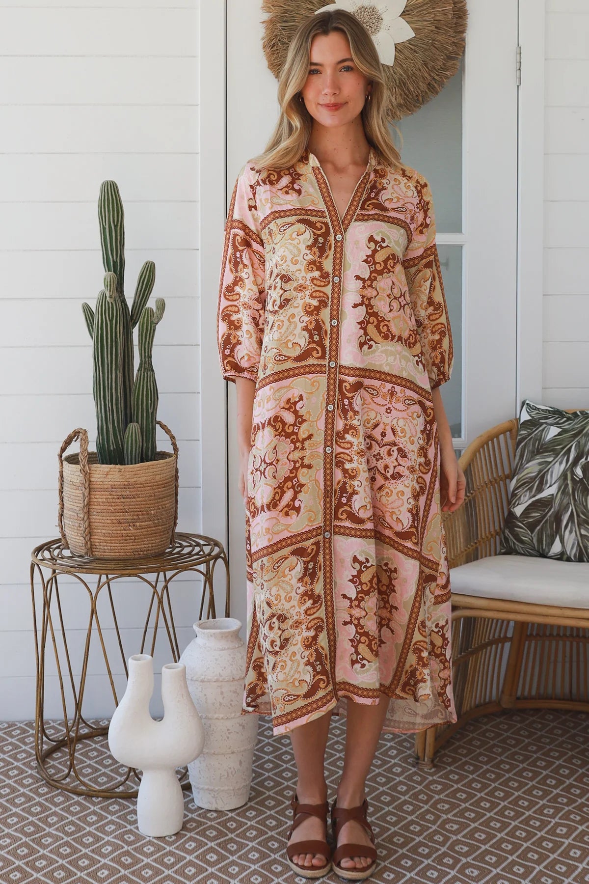 A woman stands on a porch wearing a long-sleeve bohemian-style maxi dress with a pink, brown, and beige patchwork print. She is wearing a large straw hat and brown sandals.