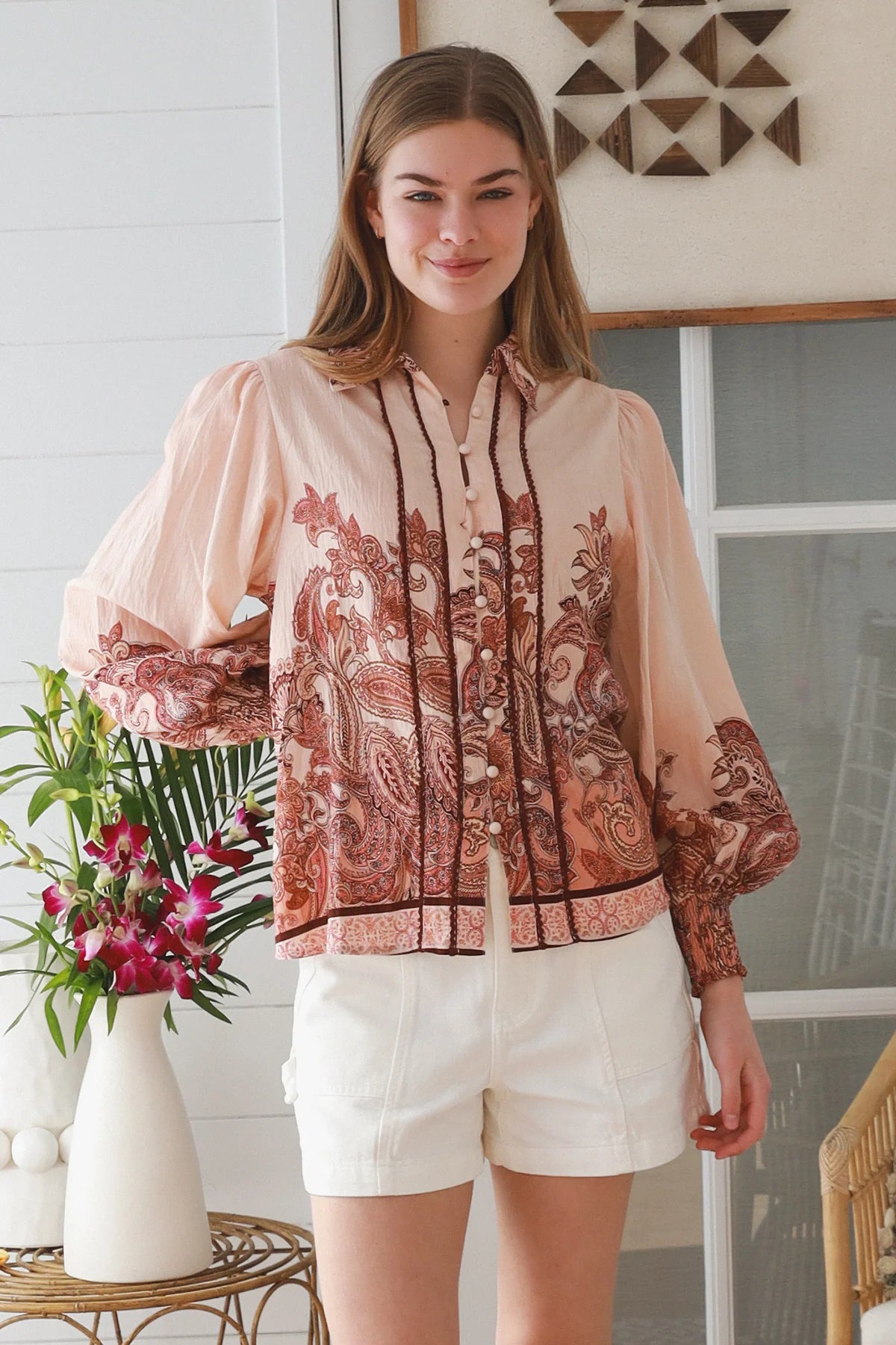 A woman modeling a sheer peach long-sleeve blouse with a brown and rust paisley print, paired with white shorts, standing in a bright room with a vase of flowers.