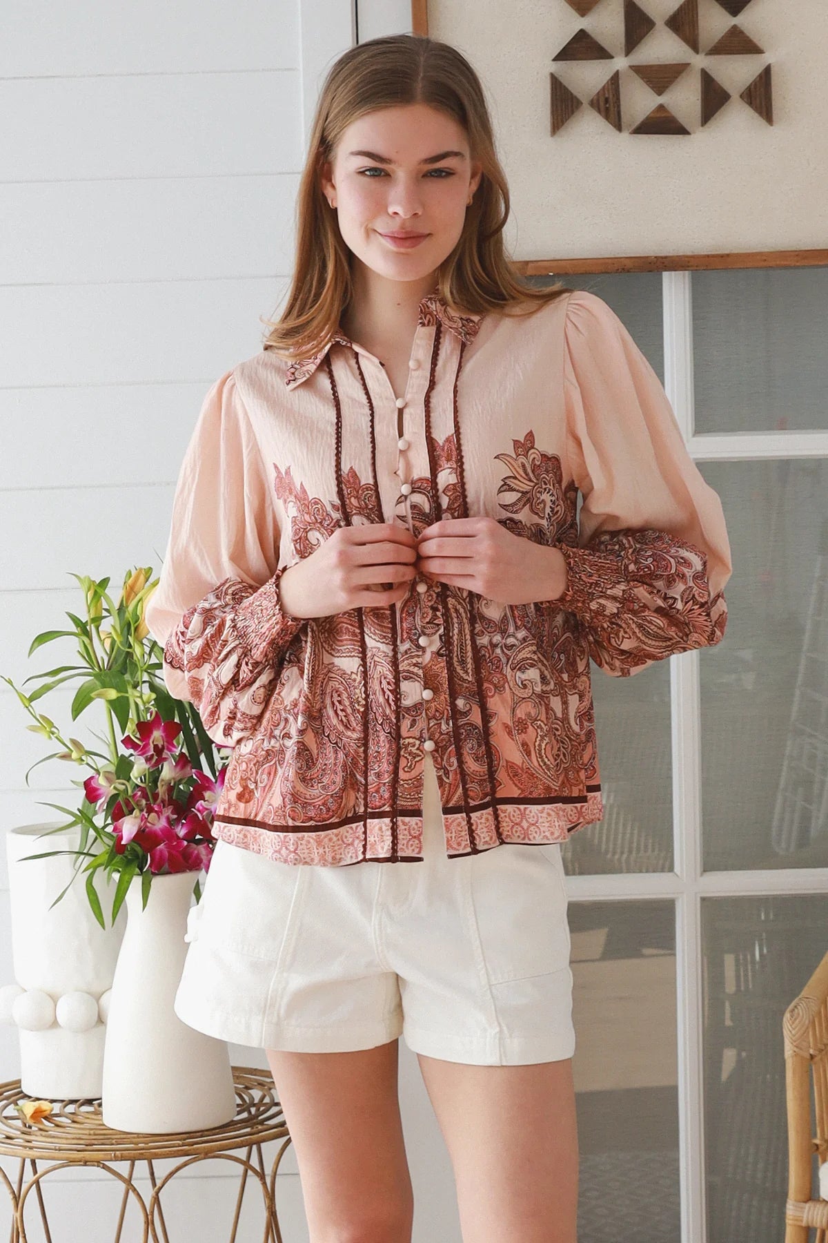 A woman wearing a peach and rust paisley print blouse with sheer balloon sleeves, paired with white shorts, standing in a bright indoor setting.