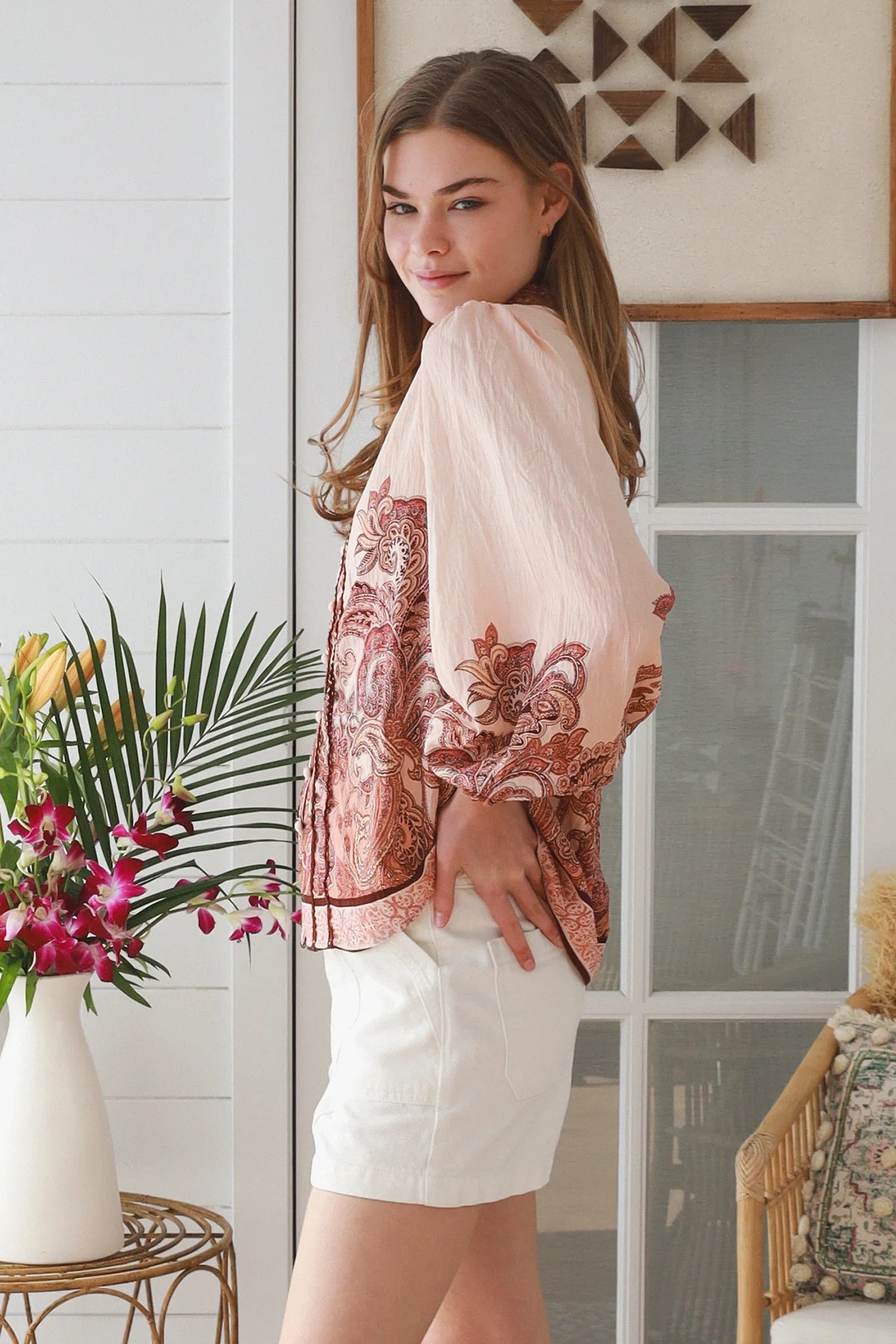 A woman wearing a sheer blush pink blouse with a rust-colored paisley pattern and white shorts, standing in a bright, modern room with a window and decorative plants.