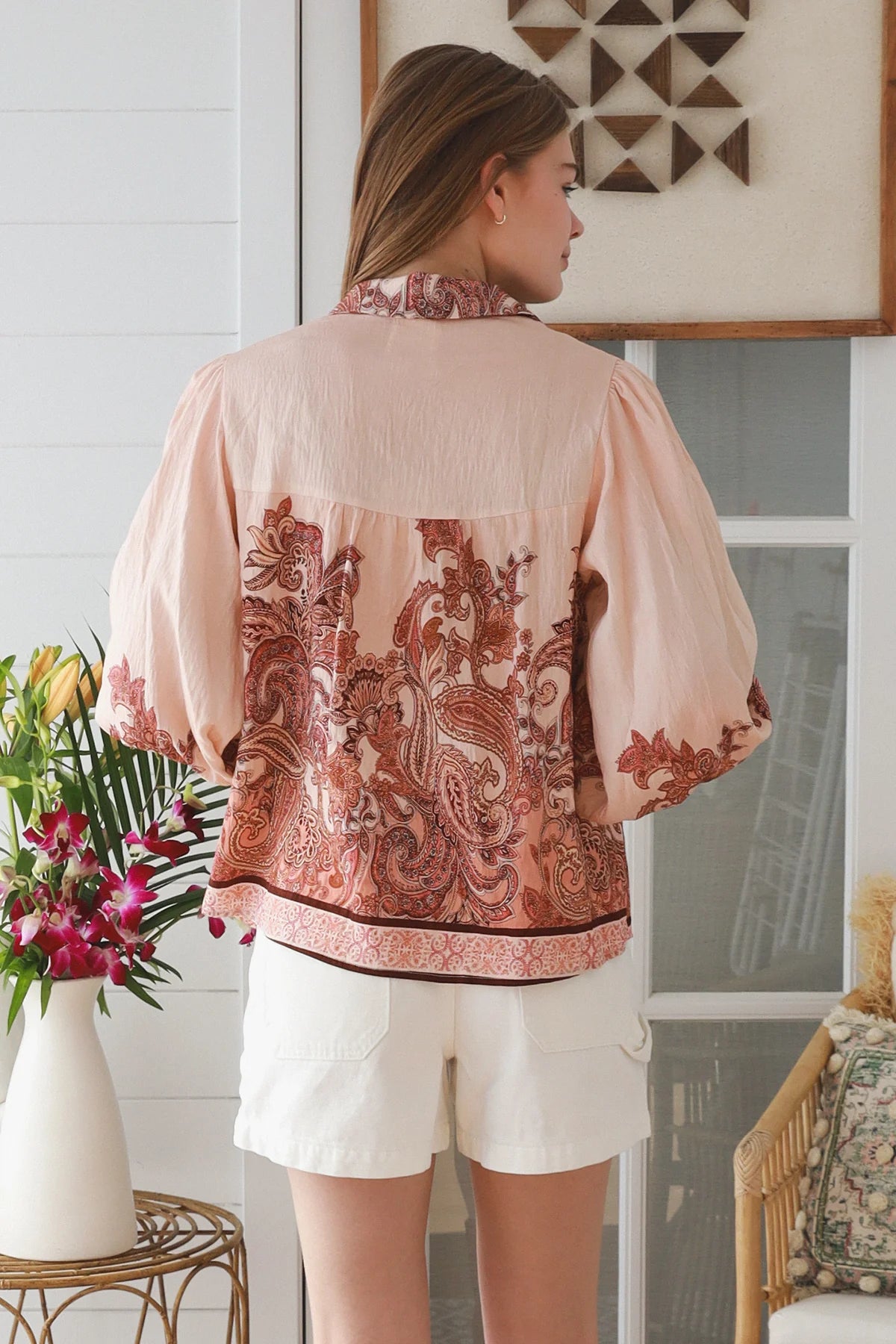 A woman shown from the back wearing a light peach pink blouse with a large rust-brown paisley pattern across the back and white shorts, standing in a bright room.