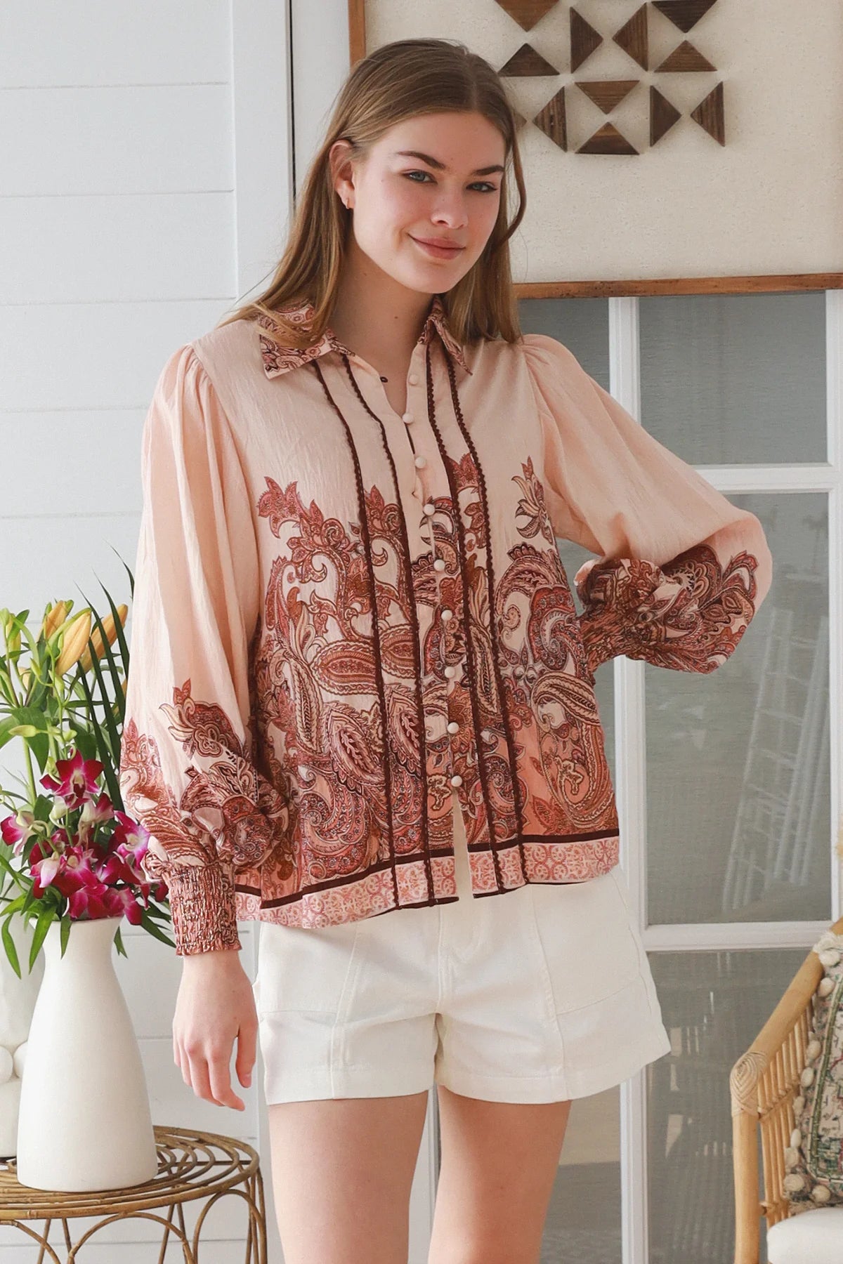 A woman modeling a peach-colored long-sleeve blouse with a brown paisley pattern and white shorts, standing in a bright, modern room with a window and decorative wall art.