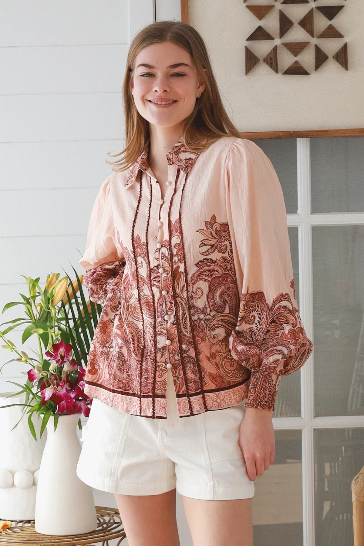 A woman wearing a light peach blouse with a rust-brown paisley pattern and white shorts, standing in a bright room with a window and decorative wall art.