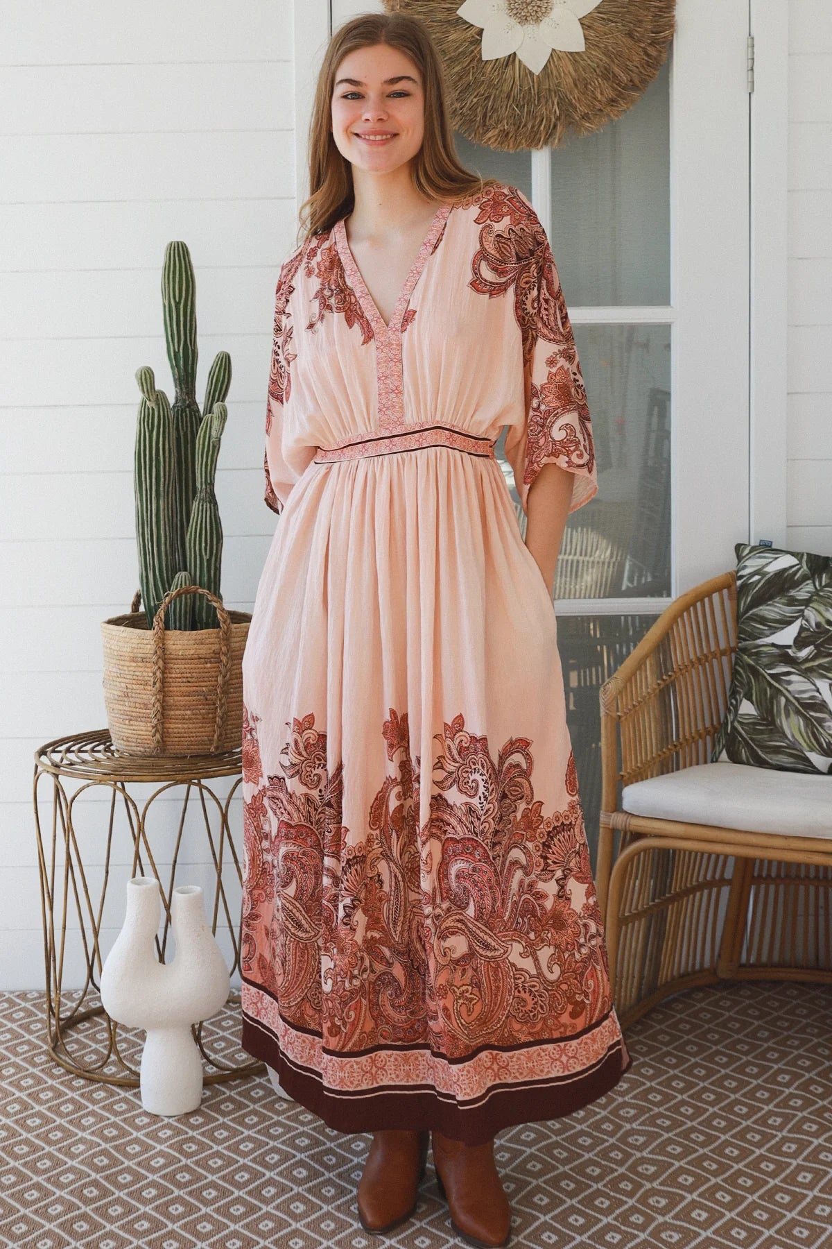 A woman wearing a peach and rust paisley maxi dress stands on a porch next to a cactus on a wire table and a woven chair.
