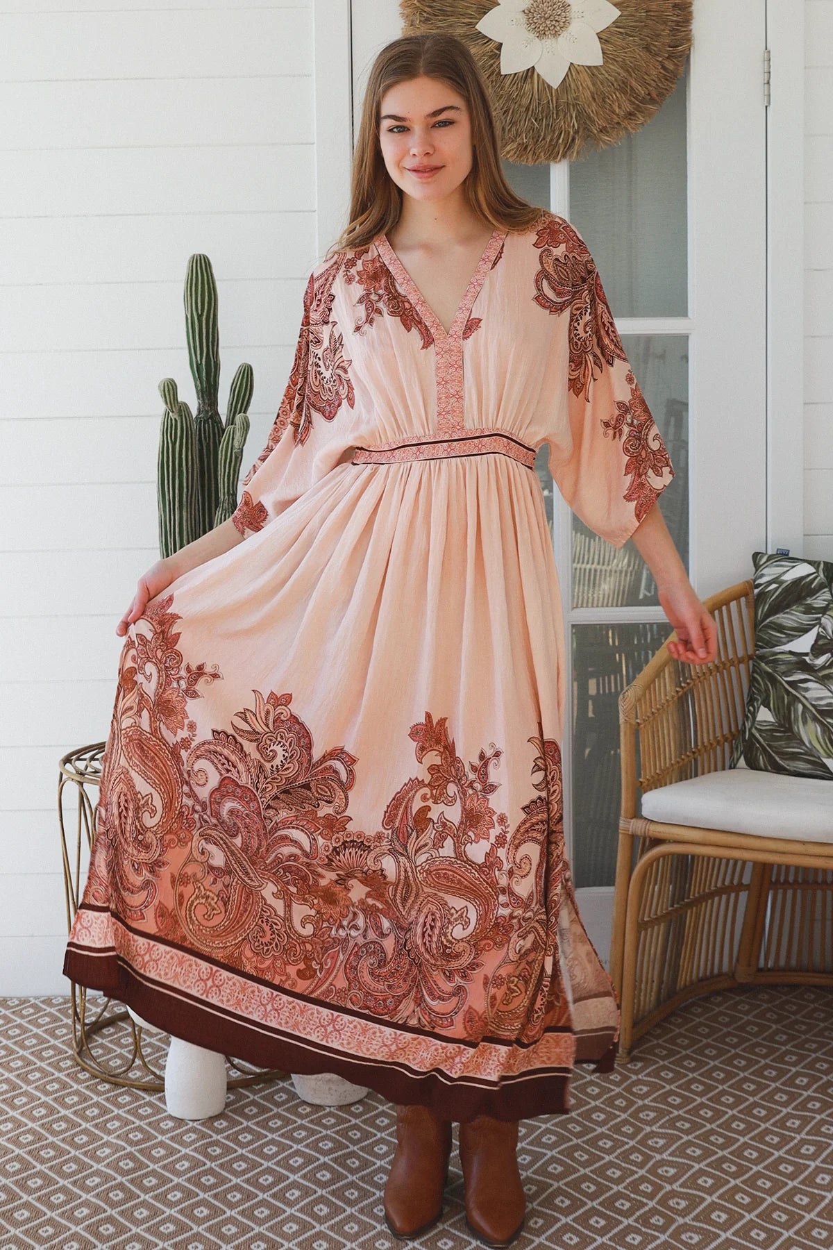 A woman models a bohemian-style maxi dress with a peach and rust paisley pattern, standing on a patterned porch floor next to a rattan chair and cactus.