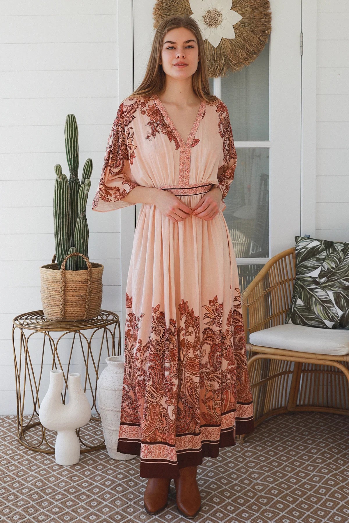 A woman stands on a porch wearing a flowing peach maxi dress with brown paisley and floral patterns, paired with brown ankle boots. The setting includes a cactus on a wire table, a wicker chair, and a white shiplap wall.