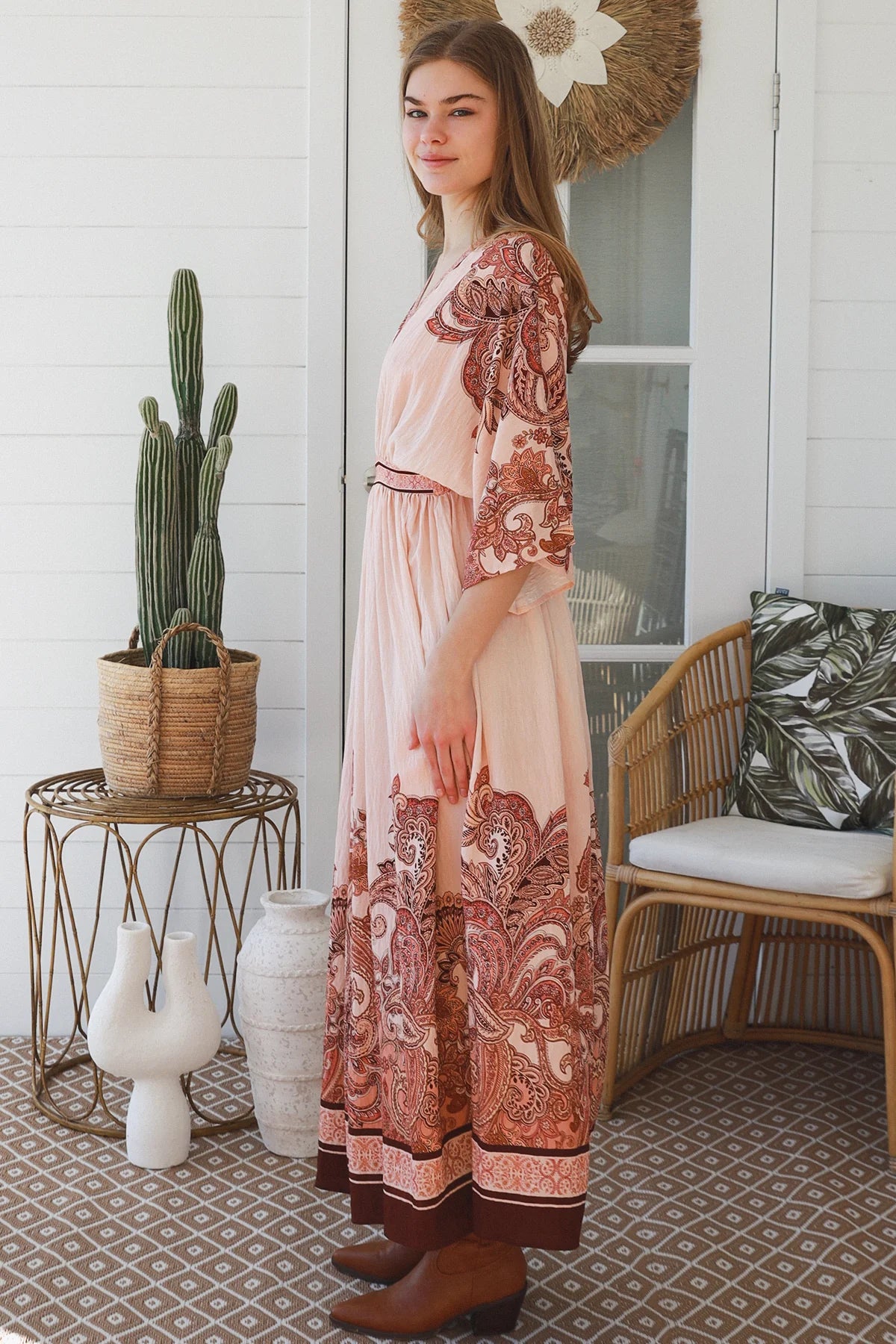 A woman stands on a porch wearing a long peach-pink maxi dress with a rust-brown paisley border and three-quarter sleeves. The setting includes a cactus on a wire side table, white ceramic vases, a wicker chair with a leaf-patterned cushion, and a white shiplap wall.