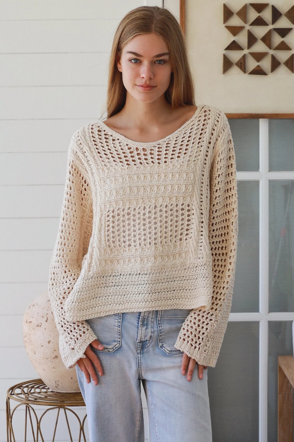 Woman wearing a beige crochet sweater and light blue jeans indoors.
