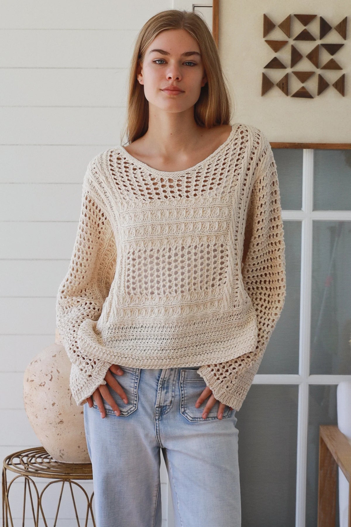 Woman wearing a beige crochet sweater and light blue jeans indoors.