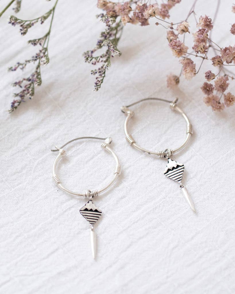 Silver hoop earrings with heart and arrow charms on a white fabric background with dried flowers.